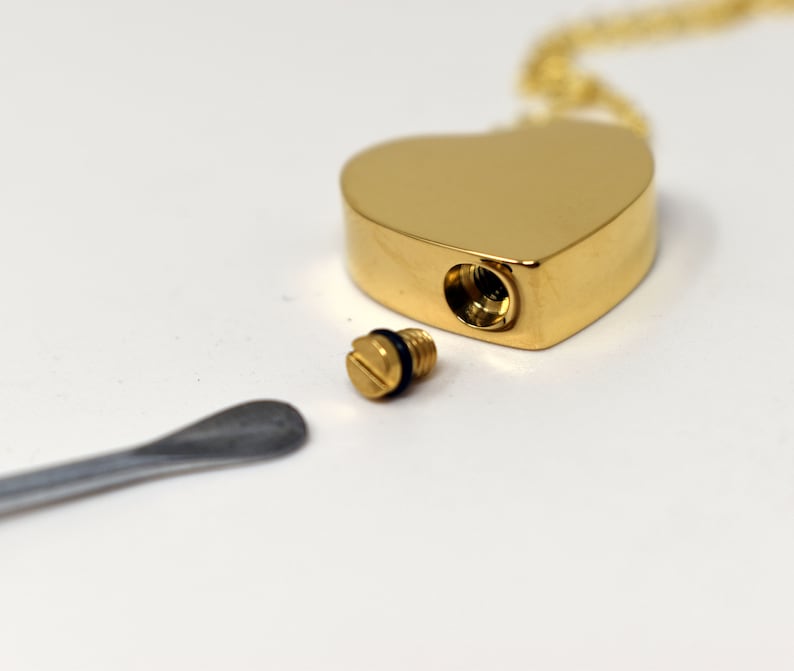 May include: A gold-colored heart-shaped pendant necklace with a screw-top opening. A small, gold-colored screw and a silver-colored spoon are next to the pendant. The pendant is on a gold chain.