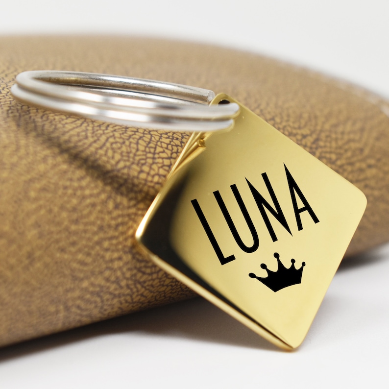 May include: Gold-colored metal keychain with a square tag that says "LUNA" in black lettering with a black crown underneath.