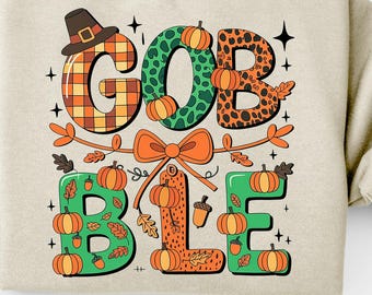 May include: Beige sweatshirt with the word "GOBBLE" in colorful, fall-themed lettering. The letters are decorated with pumpkins, leaves, and a pilgrim hat. A large orange bow and sprigs of leaves and acorns complete the design. The sweatshirt is a cozy, seasonal apparel item.
