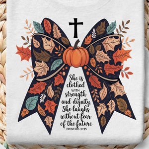 Könnte beinhalten: Weißes T-Shirt mit einer schwarzen und braunen Schleife aus Herbstblättern und einem Kürbis in der Mitte. Der Text "She is clothed with strength and dignity. She laughs without fear of the future. Proverbs 31:25" ist unter der Schleife gedruckt.