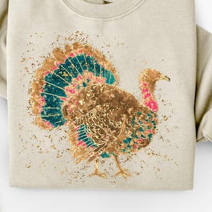 May include: A beige sweatshirt featuring a detailed illustration of a turkey with gold, teal, and pink accents. The turkey design is centered on the front of the sweatshirt, with a speckled gold background. The sweatshirt has a crew neck and long sleeves.