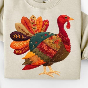 May include: A beige sweatshirt with an embroidered turkey design. The turkey has a red head and wattle, a patchwork body in shades of brown, green, and orange, and a fan of tail feathers. The design is centered on the front of the sweatshirt.
