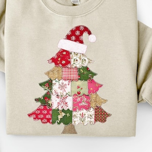 May include: Beige sweatshirt featuring a Christmas tree design. The tree is made of various fabric patches in red, green, pink, and gold, topped with a Santa hat. The design is centered on the front of the sweatshirt, perfect for the holiday season.
