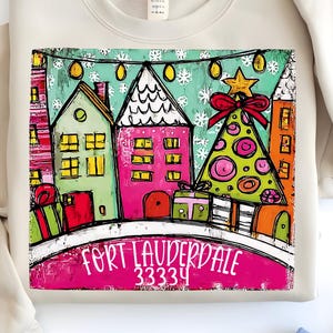 Puede incluir: Sudadera beige con una colorida ilustración de una escena navideña. El diseño incluye casas, un árbol de Navidad y el texto "FORT LAUDERDALE 33334". La obra de arte es de estilo caprichoso con colores brillantes y detalles festivos.