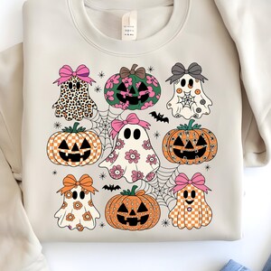 May include: Beige sweatshirt featuring a colorful Halloween-themed design. The design includes cartoon ghosts and pumpkins with bows, spiderwebs, and a bat. The ghosts have various patterns, including leopard print, floral, and checkered. The pumpkins have jack-o'-lantern faces.