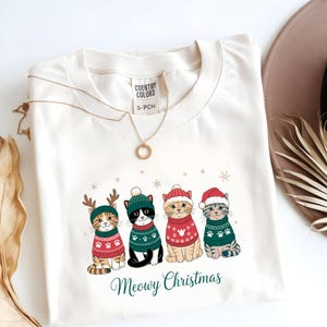 May include: Cream-colored sweatshirt featuring a festive design of four cats wearing Christmas sweaters and hats. The cats are adorned with reindeer antlers and Santa hats. The text "Meowy Christmas" is printed below the cats.