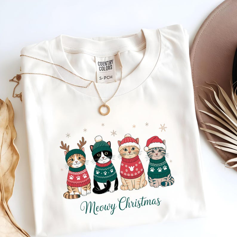 May include: Cream-colored sweatshirt featuring a festive design of four cats wearing Christmas sweaters and hats. The cats are adorned with reindeer antlers and Santa hats. The text "Meowy Christmas" is printed below the cats.
