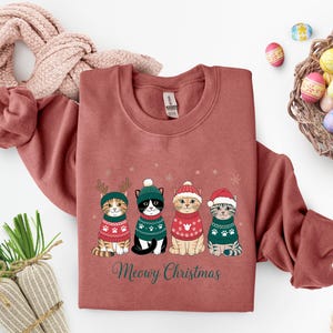 May include: A brick-red sweatshirt features four cartoon cats in festive sweaters and hats. The cats are arranged in a row, with snowflakes above them. The text "Meowy Christmas" is printed below the cats in green script.