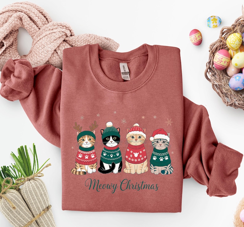 May include: A brick-red sweatshirt features four cartoon cats in festive sweaters and hats. The cats are arranged in a row, with snowflakes above them. The text "Meowy Christmas" is printed below the cats in green script.