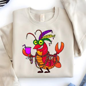 May include: Beige sweatshirt featuring a cartoon crawfish character with a festive Mardi Gras design. The crawfish is red, orange, and yellow, wearing a feathered headdress, beads, and holding a purple drink. The sweatshirt is a neutral color.