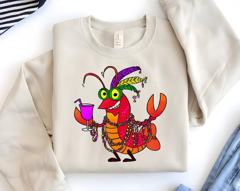 May include: Beige sweatshirt featuring a cartoon crawfish character with a festive Mardi Gras design. The crawfish is red, orange, and yellow, wearing a feathered headdress, beads, and holding a purple drink. The sweatshirt is a neutral color.