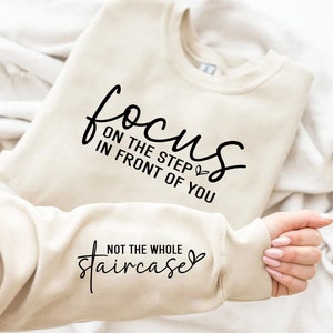 May include: A beige sweatshirt with the text "focus on the step in front of you" and "not the whole staircase" printed on it.