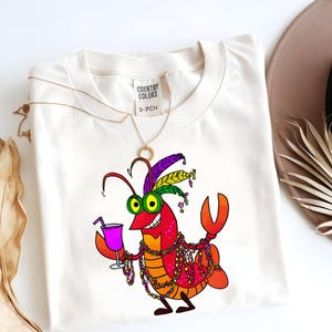 May include: Cream-colored t-shirt featuring a cartoon lobster with a cocktail, Mardi Gras beads, and a feathered headdress. The lobster is red, orange, and yellow with green eyes. The shirt has a tag that reads "COUNTRY COLORS" and "S-PCH".
