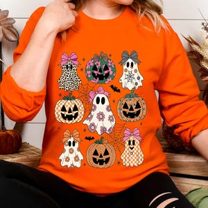 May include: Orange sweatshirt featuring a Halloween-themed design. The design includes various pumpkins and ghosts with bows, set against a spiderweb background. The pumpkins and ghosts have different patterns and colors, creating a festive and playful look.
