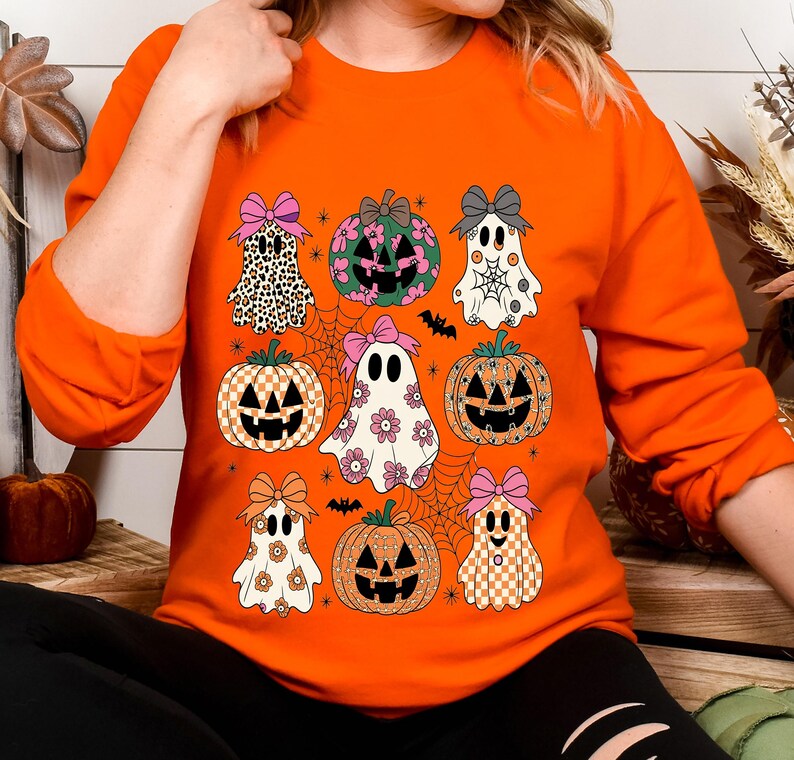 May include: Orange sweatshirt featuring a Halloween-themed design. The design includes various pumpkins and ghosts with bows, set against a spiderweb background. The pumpkins and ghosts have different patterns and colors, creating a festive and playful look.