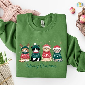 May include: Green sweatshirt featuring four cartoon cats wearing festive sweaters and hats. The cats are adorned with reindeer antlers, a beanie, a Santa hat, and a striped hat. The text "Meowy Christmas" is printed below the cats.