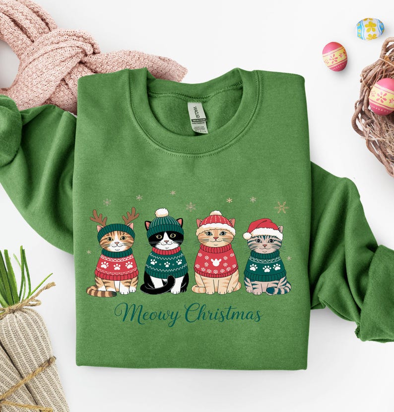 May include: Green sweatshirt featuring four cartoon cats wearing festive sweaters and hats. The cats are adorned with reindeer antlers, a beanie, a Santa hat, and a striped hat. The text "Meowy Christmas" is printed below the cats.