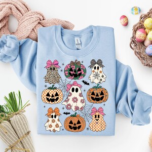 May include: Light blue sweatshirt featuring a Halloween-themed design. The design includes cartoon ghosts, pumpkins, spiderwebs, and bats. The ghosts and pumpkins have bows and various patterns, including leopard print, floral, and checkered designs. The sweatshirt is a crew neck style.