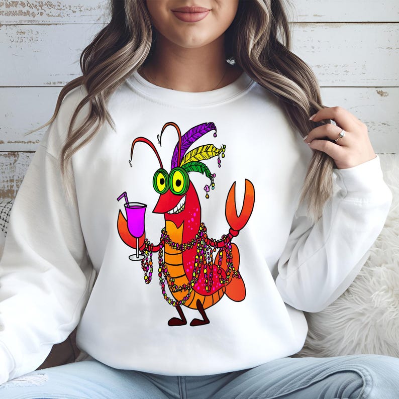 May include: White sweatshirt featuring a cartoon crawfish character with a festive Mardi Gras design. The crawfish is red, orange, and yellow, wearing a feathered headdress, beads, and holding a purple drink. The design is centered on the front of the sweatshirt.