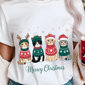 May include: White t-shirt featuring four cartoon cats in festive sweaters and hats. The cats are wearing red and green sweaters with paw print and snowflake designs. The text "Meowy Christmas" is written in green script below the cats.