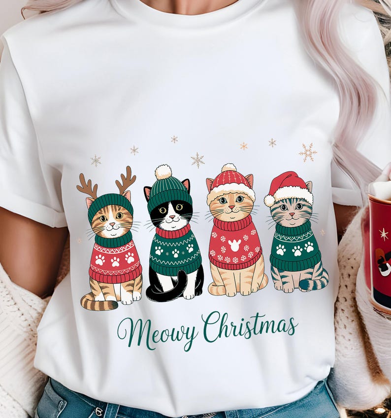 May include: White t-shirt featuring four cartoon cats in festive sweaters and hats. The cats are wearing red and green sweaters with paw print and snowflake designs. The text "Meowy Christmas" is written in green script below the cats.