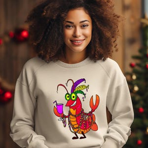 May include: A beige sweatshirt featuring a cartoon crawfish character. The crawfish is depicted with a festive Mardi Gras theme, wearing a feathered hat, beads, and holding a cocktail. The design is colorful and playful.