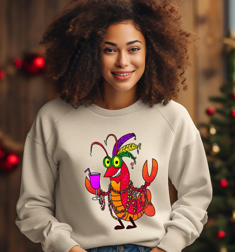 May include: A beige sweatshirt featuring a cartoon crawfish character. The crawfish is depicted with a festive Mardi Gras theme, wearing a feathered hat, beads, and holding a cocktail. The design is colorful and playful.