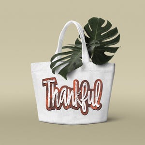 K&ouml;nnte beinhalten: Wei&szlig;e Canvas-Tragetasche mit einer braunen und wei&szlig;en "Thankful"-Grafik. Die Tasche hat ein gr&uuml;nes Blatt oben drauf.