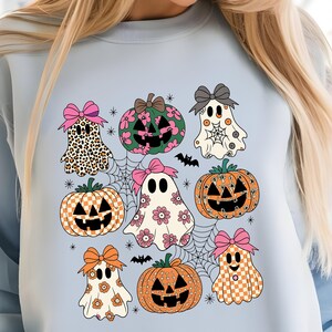 May include: Light blue sweatshirt featuring a Halloween-themed design. The design includes cartoon ghosts, pumpkins, spiderwebs, and bats. The ghosts have various patterns, including leopard print, floral, and checkered. The pumpkins have carved faces.