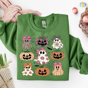 May include: Green sweatshirt featuring a Halloween-themed design with ghosts, pumpkins, and spiderwebs. The ghosts and pumpkins have bows and various patterns. The design includes bats and floral accents, perfect for the spooky season.