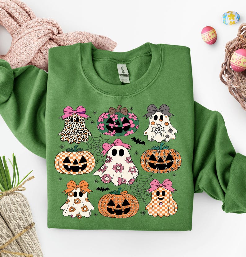 May include: Green sweatshirt featuring a Halloween-themed design with ghosts, pumpkins, and spiderwebs. The ghosts and pumpkins have bows and various patterns. The design includes bats and floral accents, perfect for the spooky season.