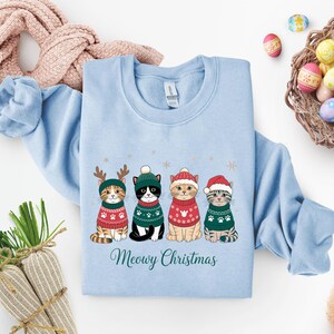 May include: Light blue sweatshirt featuring four cartoon cats in Christmas sweaters and hats. The cats are wearing red and green sweaters with paw prints. The text "Meowy Christmas" is written in a script font below the cats.