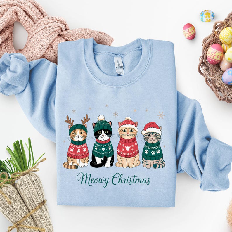 May include: Light blue sweatshirt featuring four cartoon cats in Christmas sweaters and hats. The cats are wearing red and green sweaters with paw prints. The text "Meowy Christmas" is written in a script font below the cats.