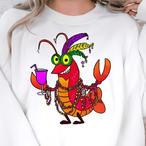 May include: White sweatshirt featuring a cartoon lobster with a festive design. The lobster is red, orange, and purple, wearing a feathered headdress, beads, and holding a purple cocktail. A fun and whimsical design.
