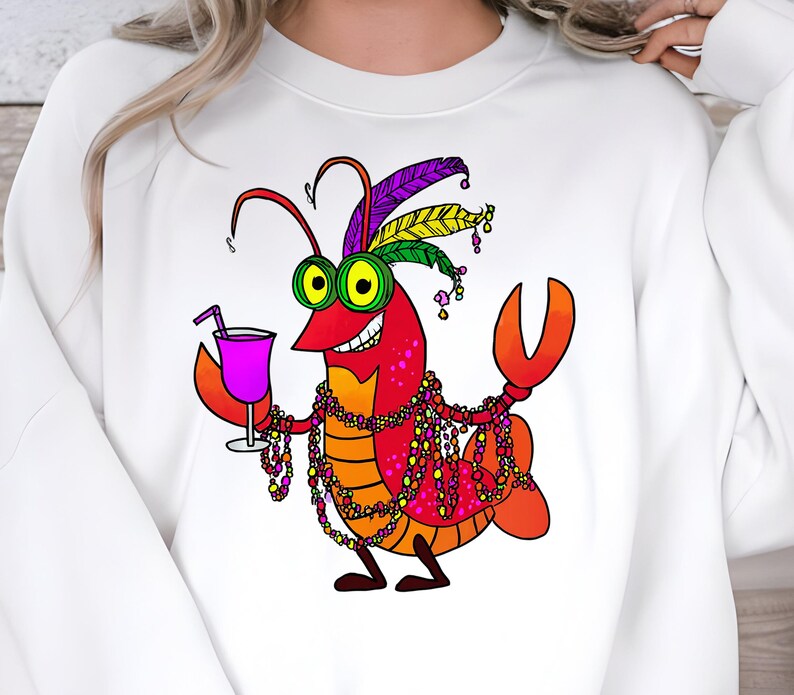 May include: White sweatshirt featuring a cartoon lobster with a festive design. The lobster is red, orange, and purple, wearing a feathered headdress, beads, and holding a purple cocktail. A fun and whimsical design.