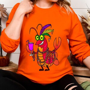 May include: Orange long-sleeve sweatshirt featuring a cartoon crawfish character with green eyes, holding a pink cocktail. The crawfish is adorned with a feathered headdress and beaded necklaces. The design is colorful and festive.