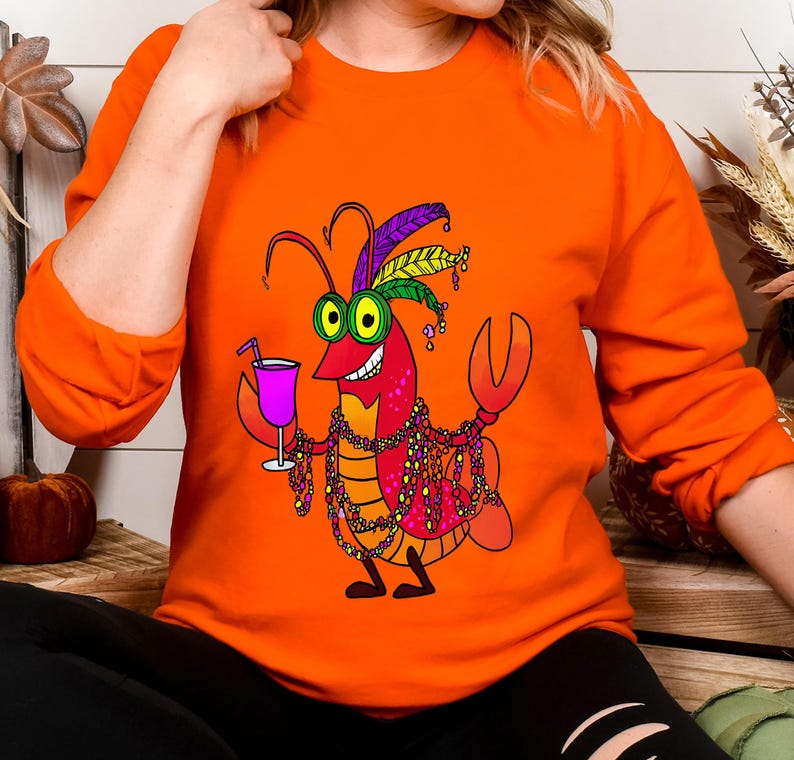 May include: Orange long-sleeve sweatshirt featuring a cartoon crawfish character with green eyes, holding a pink cocktail. The crawfish is adorned with a feathered headdress and beaded necklaces. The design is colorful and festive.