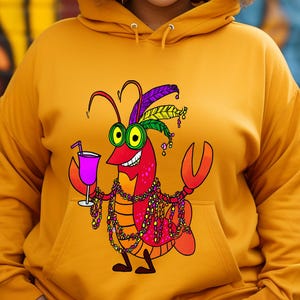 May include: Mustard yellow hoodie featuring a cartoon crawfish with Mardi Gras beads, a feathered headdress, and a cocktail. The crawfish is red with yellow eyes and a big smile. The hoodie has a drawstring hood and a front pocket.