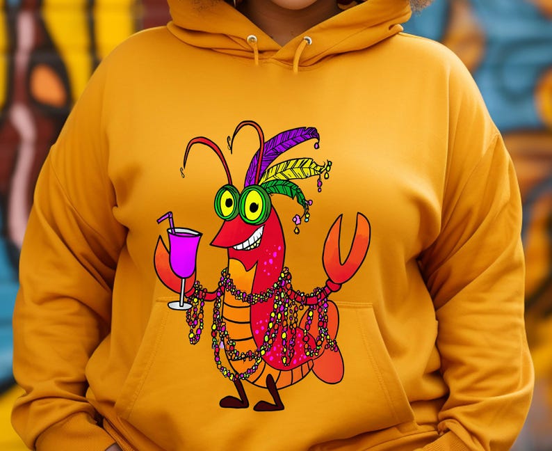May include: Mustard yellow hoodie featuring a cartoon crawfish with Mardi Gras beads, a feathered headdress, and a cocktail. The crawfish is red with yellow eyes and a big smile. The hoodie has a drawstring hood and a front pocket.
