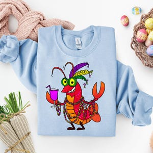 May include: Light blue sweatshirt featuring a cartoon lobster with a festive design. The lobster is red and orange, wearing a feathered headdress, beads, and holding a pink cocktail. The design is centered on the front of the sweatshirt.