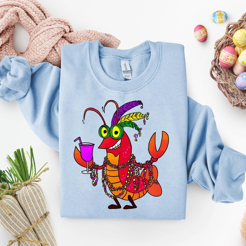 May include: Light blue sweatshirt featuring a cartoon lobster with a festive design. The lobster is red and orange, wearing a feathered headdress, beads, and holding a pink cocktail. The design is centered on the front of the sweatshirt.
