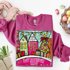 May include: A magenta sweatshirt featuring a colorful graphic of houses, a Christmas tree, and gifts. The graphic includes the text "FORT LAUDERDALE 33334". The sweatshirt is surrounded by Easter eggs and a scarf.