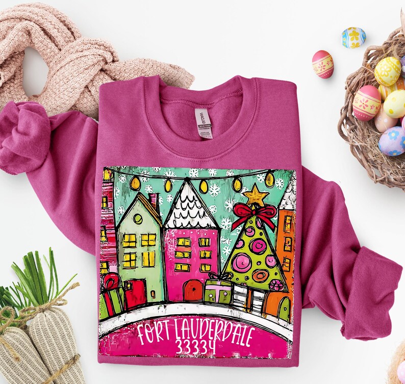 May include: A magenta sweatshirt featuring a colorful graphic of houses, a Christmas tree, and gifts. The graphic includes the text "FORT LAUDERDALE 33334". The sweatshirt is surrounded by Easter eggs and a scarf.