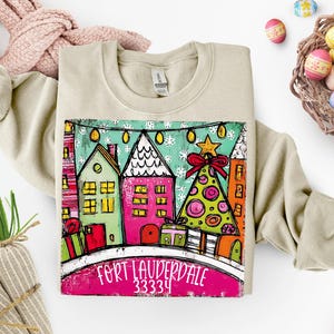 May include: Beige sweatshirt featuring a colorful graphic of houses and a Christmas tree. The graphic includes the text "Fort Lauderdale 33334". The design has a whimsical, hand-painted style with bright colors and festive elements.