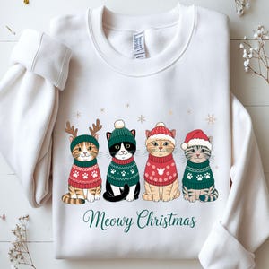 May include: White sweatshirt featuring four cartoon cats dressed in Christmas sweaters and hats. The cats are wearing green and red sweaters with paw print designs. The text "Meowy Christmas" is written in green script below the cats.