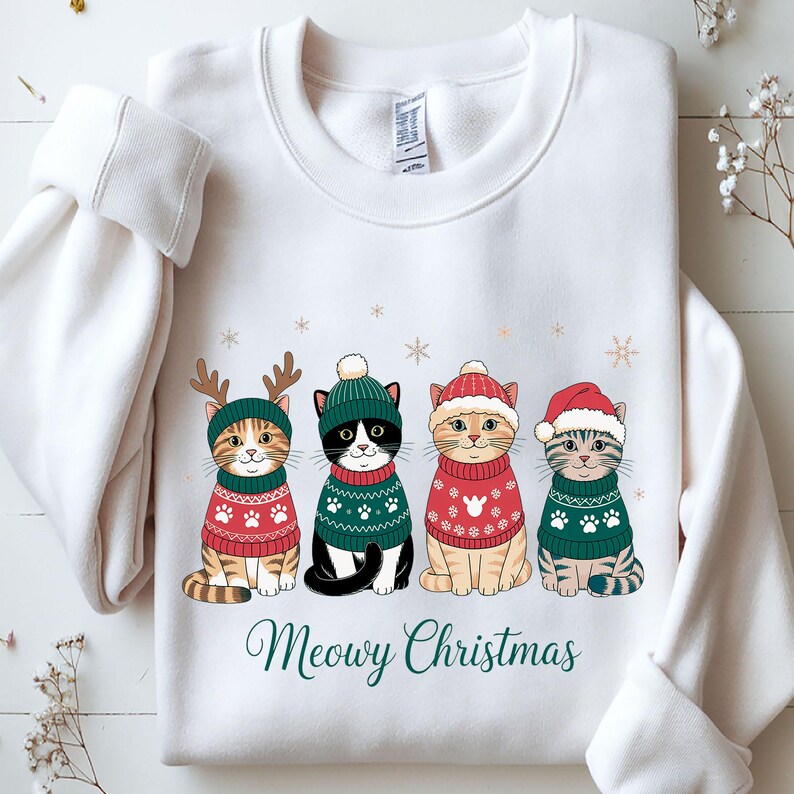 May include: White sweatshirt featuring four cartoon cats dressed in Christmas sweaters and hats. The cats are wearing green and red sweaters with paw print designs. The text "Meowy Christmas" is written in green script below the cats.