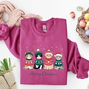 May include: A magenta sweatshirt featuring four cartoon cats in festive sweaters and hats. The cats are arranged in a row, with the text "Meowy Christmas" below. The design includes snowflakes and reindeer antlers.