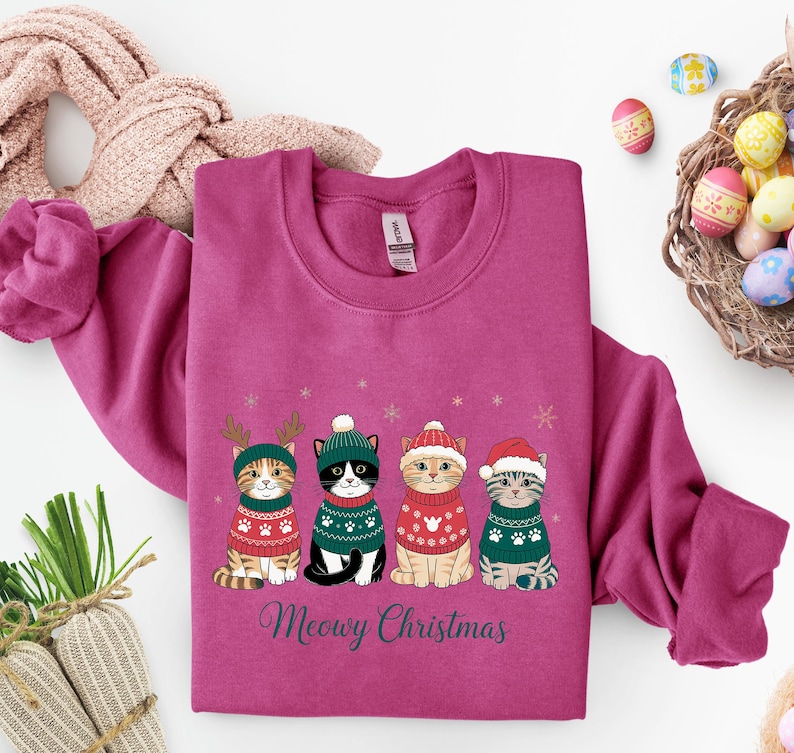 May include: A magenta sweatshirt featuring four cartoon cats in festive sweaters and hats. The cats are arranged in a row, with the text "Meowy Christmas" below. The design includes snowflakes and reindeer antlers.