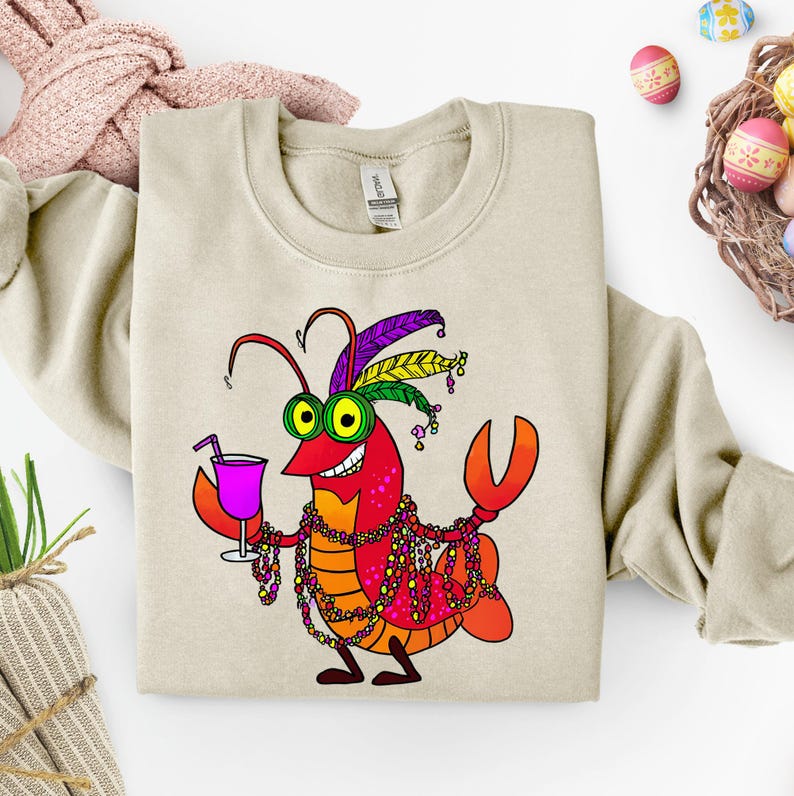 May include: Beige sweatshirt featuring a cartoon crawfish with a festive Mardi Gras design. The crawfish is red, orange, and yellow, wearing a feathered headdress, beads, and holding a purple drink. The design is centered on the front of the sweatshirt.