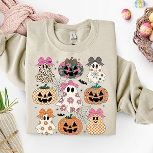 May include: Beige sweatshirt featuring a Halloween-themed design. The design includes cartoon ghosts, pumpkins, spiderwebs, and bats. The ghosts and pumpkins have bows and various patterns. The overall color palette includes orange, pink, black, and white.